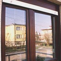 SQL biuro obsługi inwestycji - Brązowe okno PCV z roletą zewnętrzną i widokiem na budynek mieszkalny odbijający się w szybie.